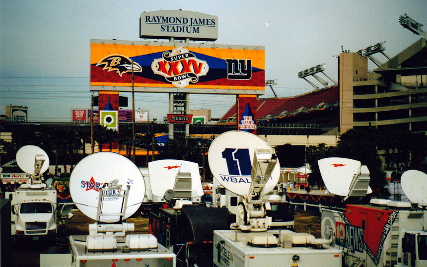 A row of satellite trucks and dishes outside a stadium under a Super Bowl XXXV scoreboard banner