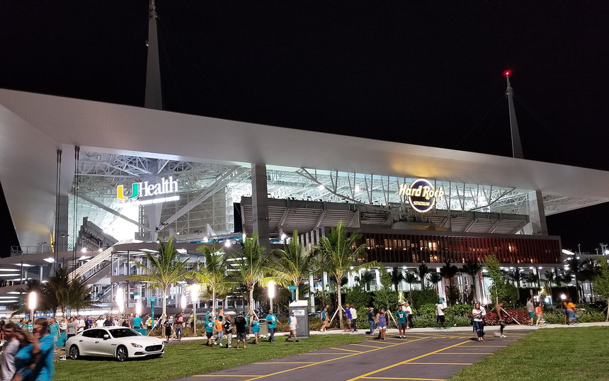 The canopy and lit concourse of Hard Rock Stadium at dusk, palms and parked cars in front