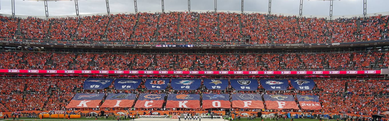 A packed NFL stadium with KICKOFF banners along the lower deck and flags laid out across the field before the game