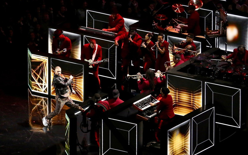 A performer on a darkened field stage of mirrored frames, a band in red playing behind him