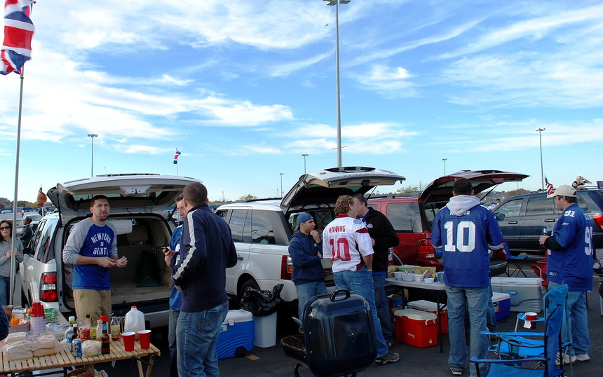 Fans in jerseys around open car hatches with coolers and folding chairs in a stadium parking lot