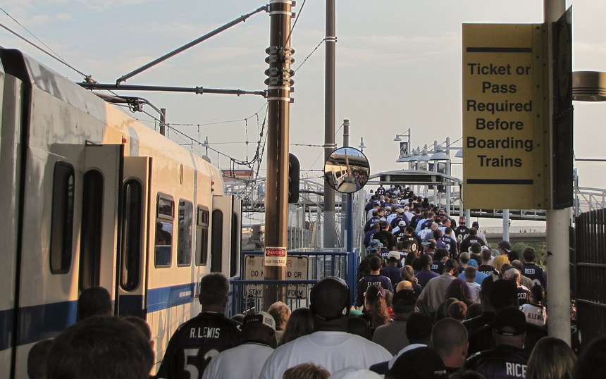 A crowded light-rail platform of fans in jerseys beside a sign reading Ticket or Pass Required Before Boarding Trains